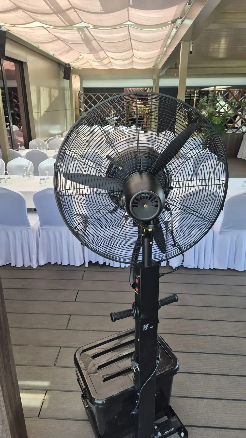 Ventilator s maglicom na terasi restorana postavljenoj za proslavu, uz stolove prekrivene bijelim stolnjacima